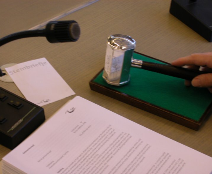 De voorzittershamer van de Gemeenraad Oss die wordt geslagen op een blok. Daarnaast ligt een stembriefje en een pak papier waarop iets is getypt